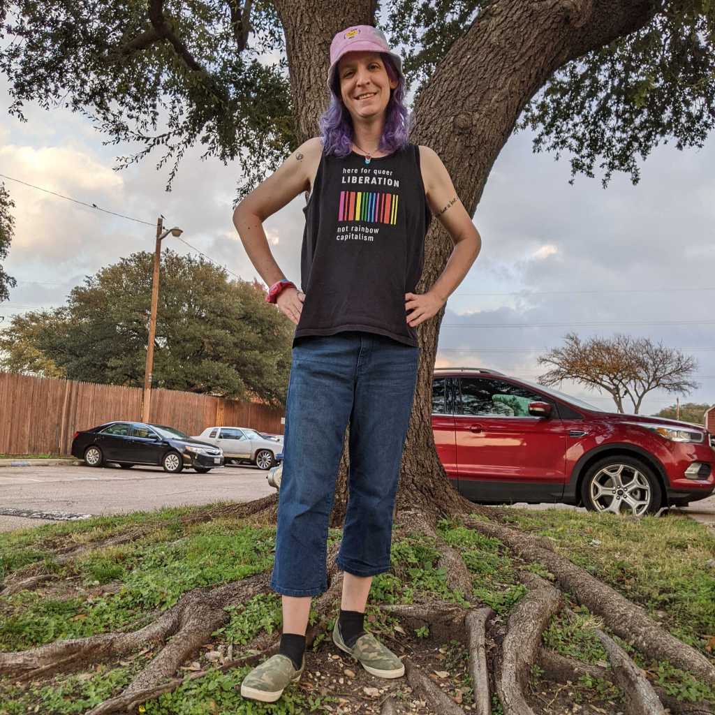 Me, a trans woman with shoulder-length purple hair, wearing a hat that says "Protect Trans Kids" and a shirt reading "Here for Queer Liberation, Not Rainbow Capitalism"